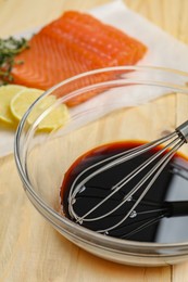 Photo of Soy sauce in bowl, whisk, salmon fillet and lemon slices on wooden table, closeup