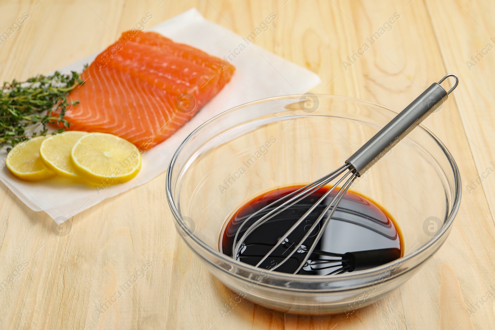 Photo of Soy sauce in bowl, whisk, salmon fillet, thyme and lemon slices on wooden table, closeup