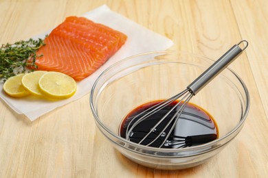 Photo of Soy sauce in bowl, whisk, salmon fillet, thyme and lemon slices on wooden table, closeup