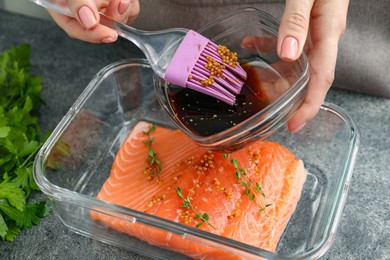 Photo of Woman marinating salmon fillet in dish at gray textured table, closeup