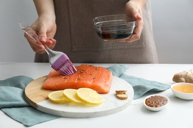 Photo of Woman marinating salmon fillet in dish at white table, closeup