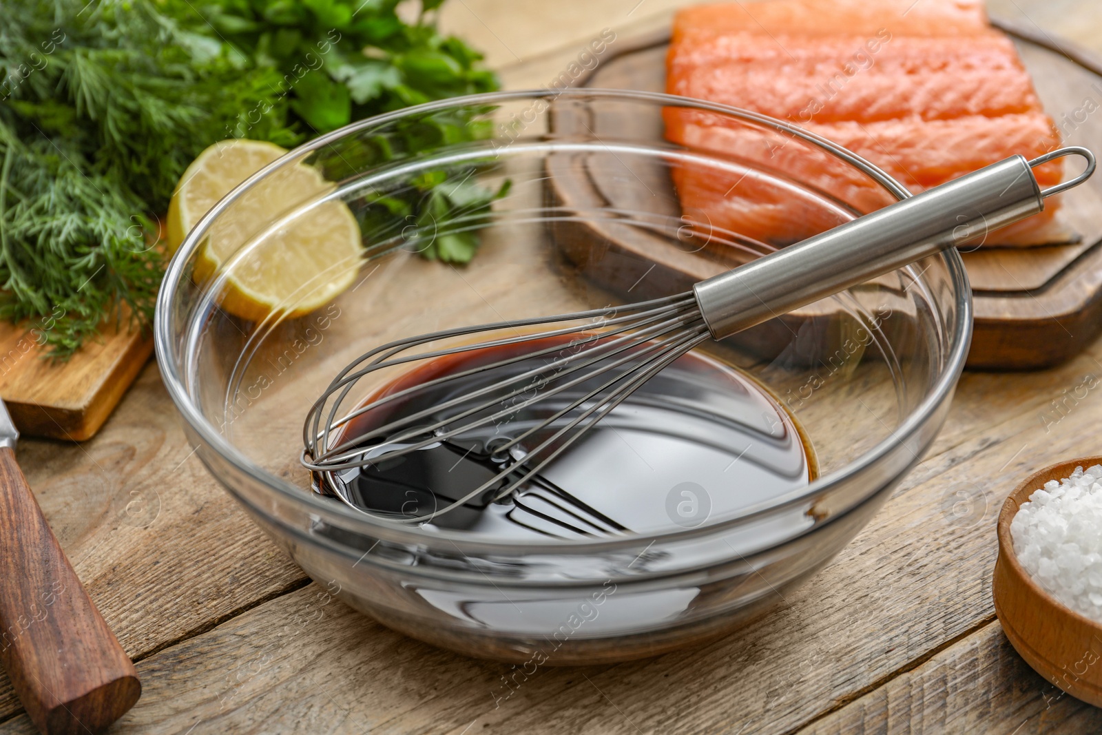 Photo of Soy sauce in bowl, whisk, salmon fillet, herbs and lemon on wooden table, closeup