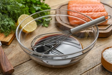 Photo of Soy sauce in bowl, whisk, salmon fillet, herbs and lemon on wooden table, closeup