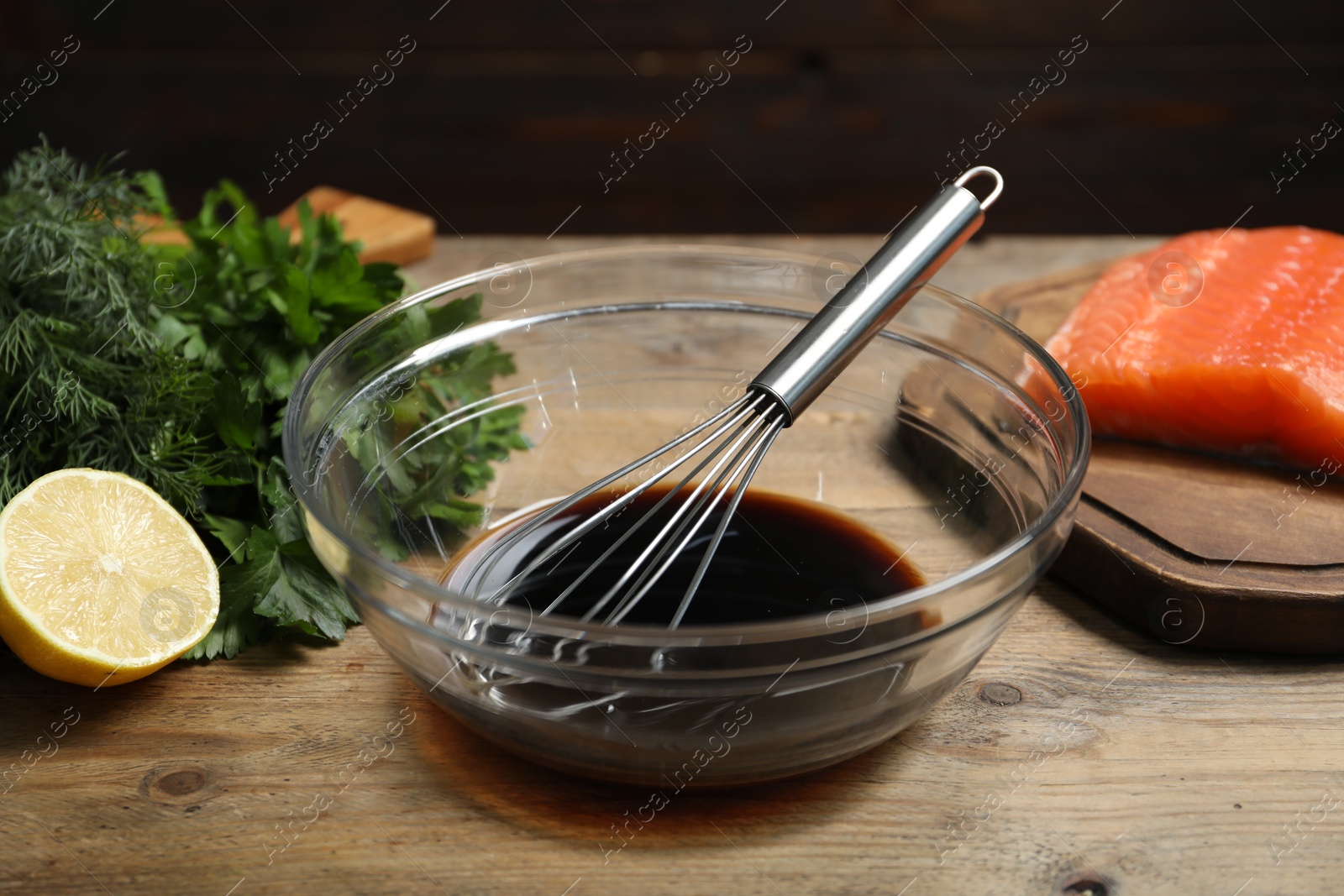 Photo of Soy sauce in bowl, whisk, salmon fillet, herbs and lemon on wooden table, closeup