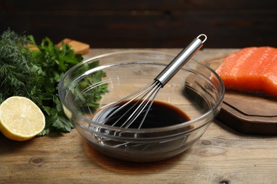 Photo of Soy sauce in bowl, whisk, salmon fillet, herbs and lemon on wooden table, closeup