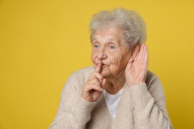 Senior woman showing hand to ear gesture on dark yellow background