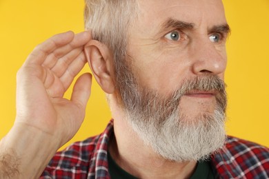 Senior man showing hand to ear gesture on orange background, closeup