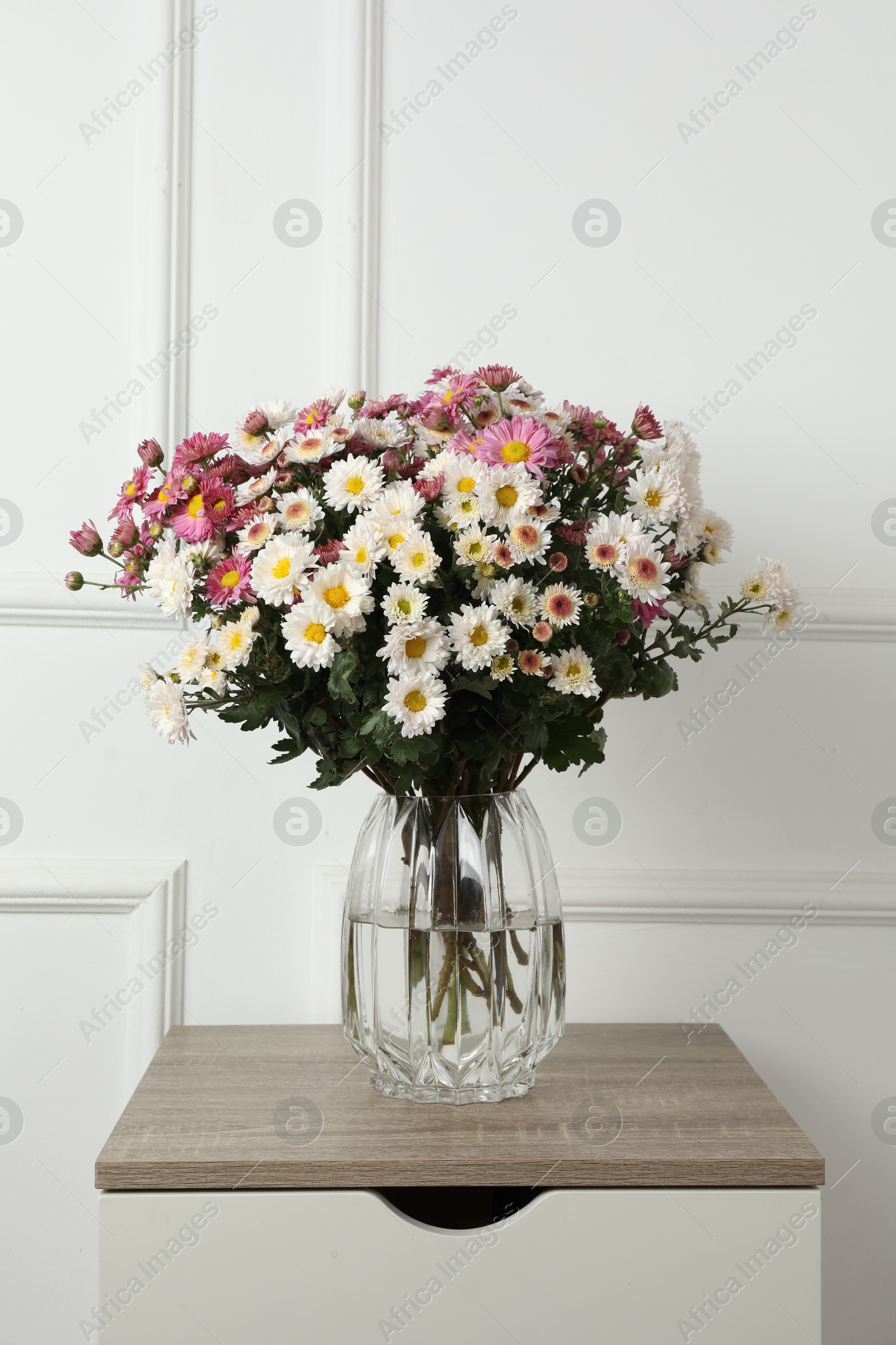 Photo of Vase with beautiful flowers on wooden nightstand near white wall