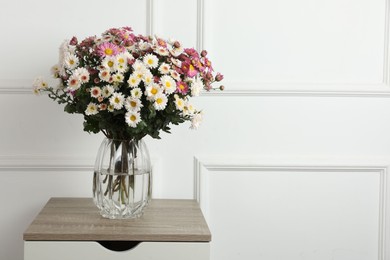 Photo of Vase with beautiful flowers on wooden nightstand near white wall, space for text