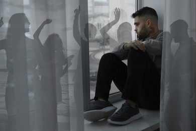 Loneliness. Sad man sitting near window in room and group of friends having fun outdoors, double exposure