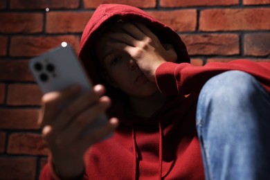 Loneliness concept. Sad teenage boy with smartphone near brick wall, selective focus