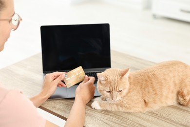 Online pet shop. Woman with laptop, credit card and cute cat making order at wooden table indoors, closeup