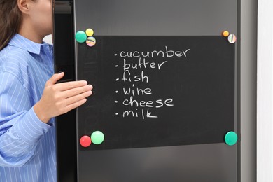 Photo of Woman opening refrigerator with shopping list on magnetic board in kitchen, closeup