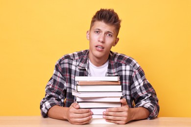 Young student with stack of books having stress before exam at wooden table against yellow background