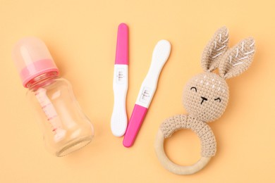 Photo of Pregnancy tests, rattle and baby bottle on pale orange background, flat lay