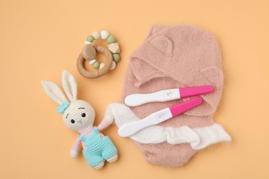 Photo of Pregnancy tests, rattle, toy bunny and baby clothes on pale orange background, top view