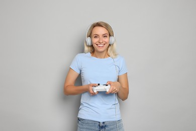 Happy woman in headphones playing video games with controller on gray background