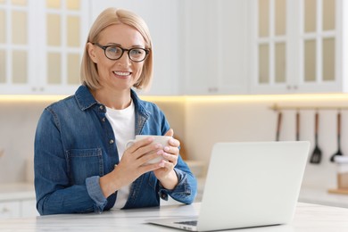 Photo of Happy middle aged woman with cup of drink using laptop at white marble table in kitchen