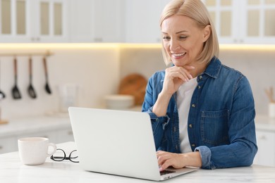 Happy middle aged woman using laptop at white marble table in kitchen