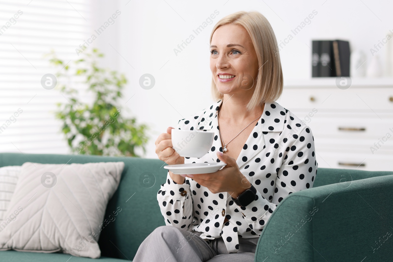 Photo of Smiling middle aged woman with cup of hot drink on sofa at home. Space for text