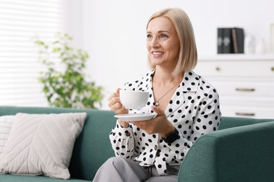 Photo of Smiling middle aged woman with cup of hot drink on sofa at home. Space for text