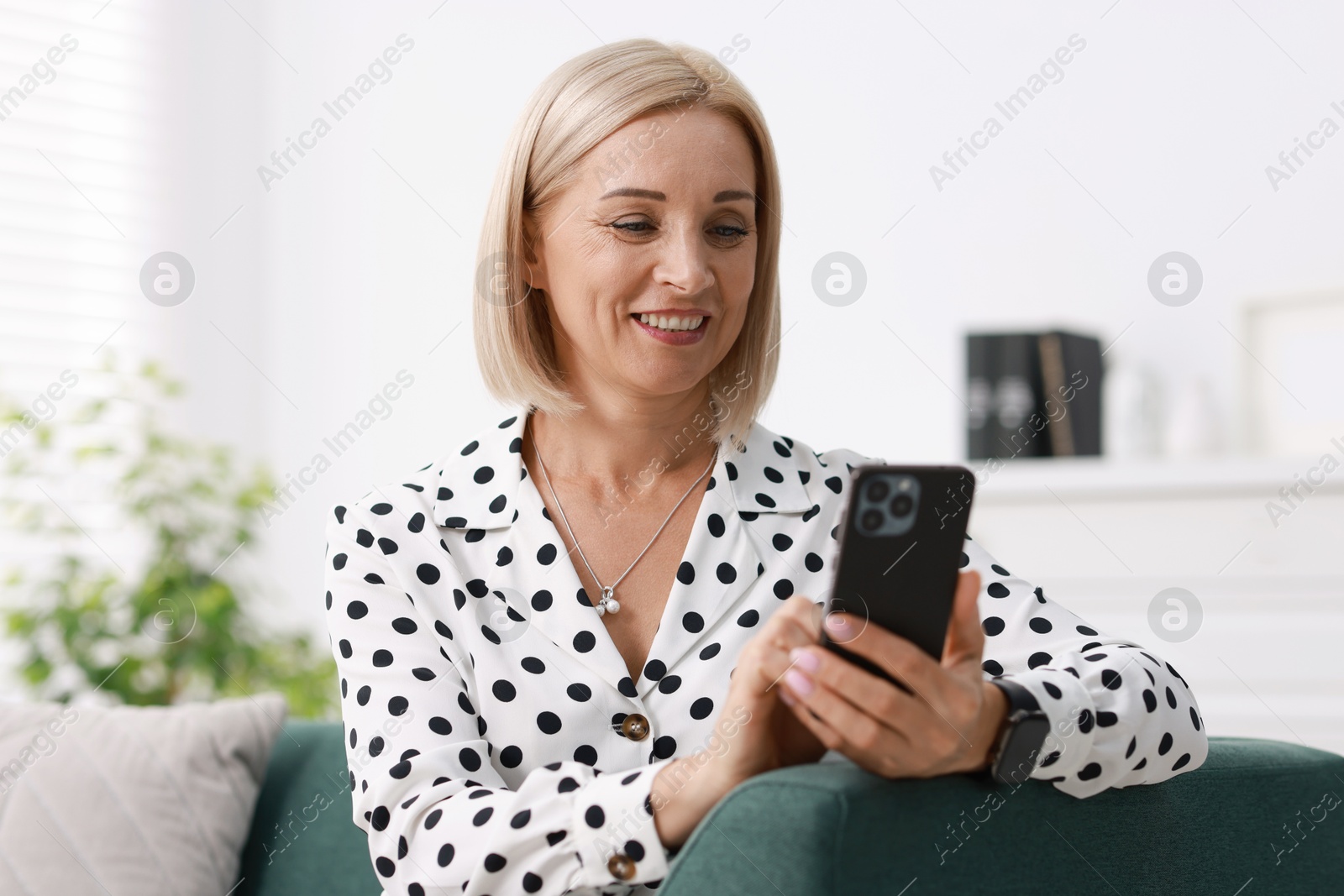 Photo of Portrait of smiling middle aged woman using smartphone on sofa at home