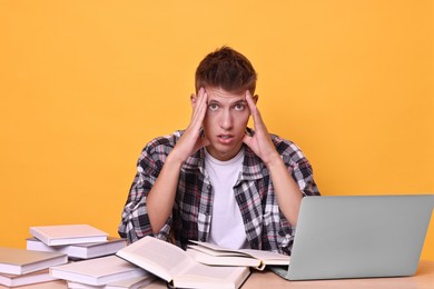 Young student with books and laptop having stress before exam at table against yellow background