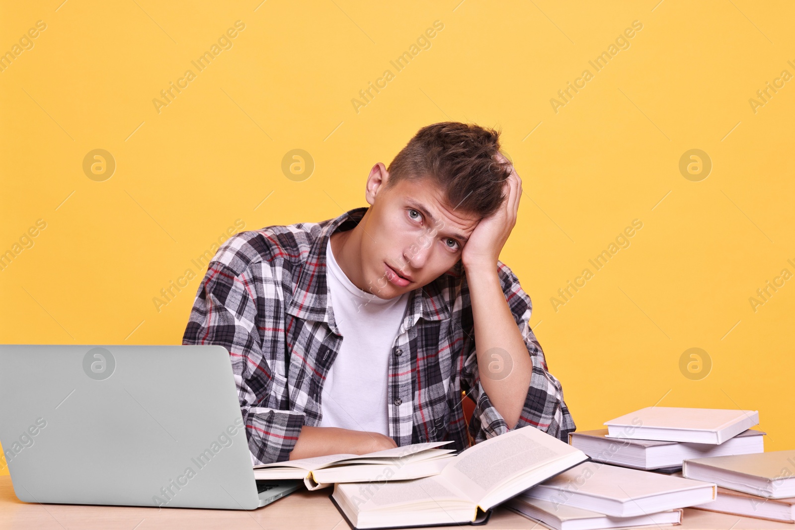 Photo of Young student with books and laptop having stress before exam at table against yellow background