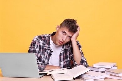 Photo of Young student with books and laptop having stress before exam at table against yellow background