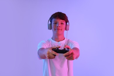 Teenage boy in headphones playing video game with controller on violet background