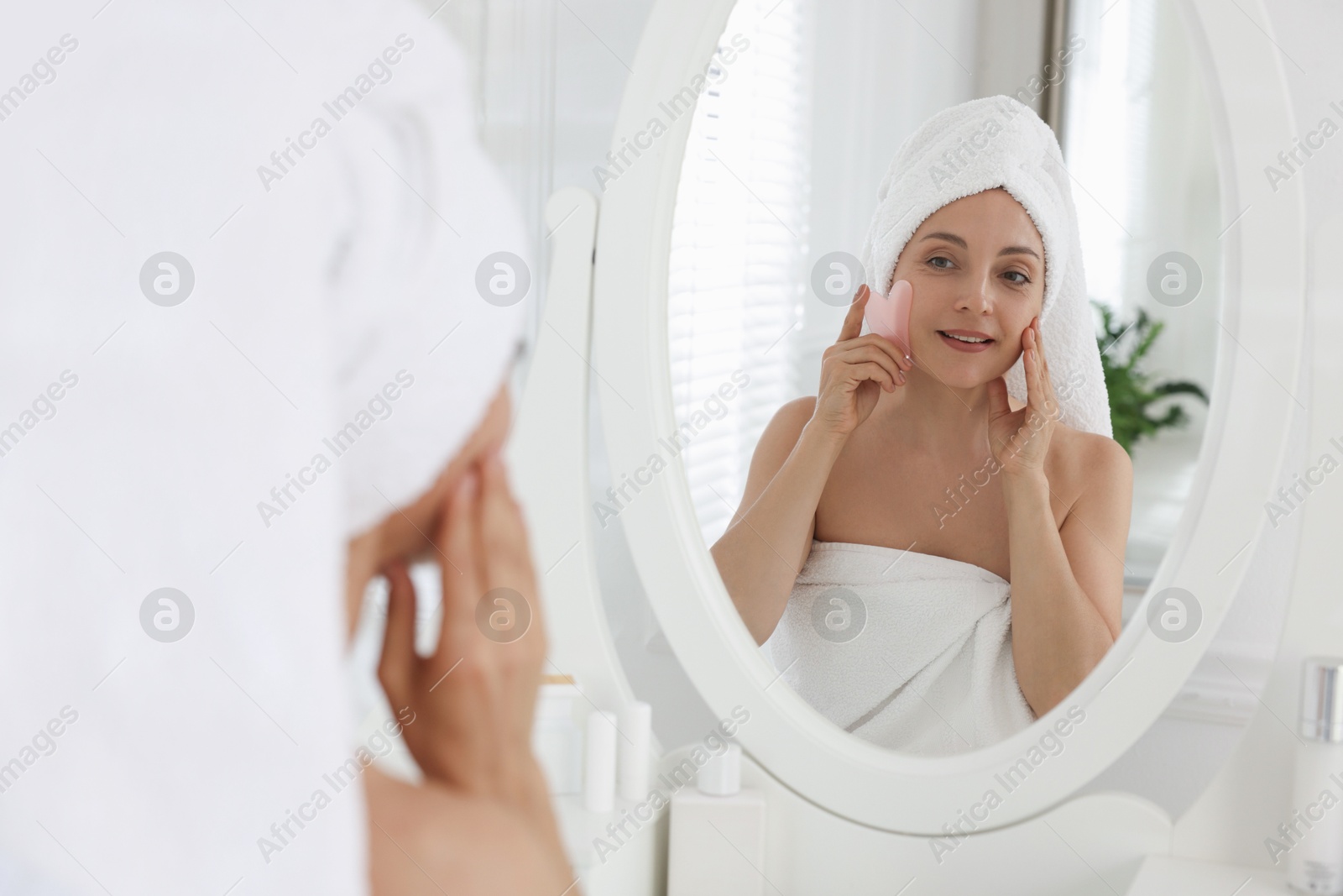 Photo of Smiling woman doing facial self massage with gua sha tool near mirror in bathroom