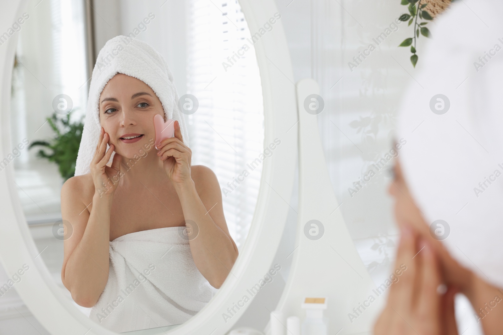 Photo of Smiling woman doing facial self massage with gua sha tool near mirror in bathroom