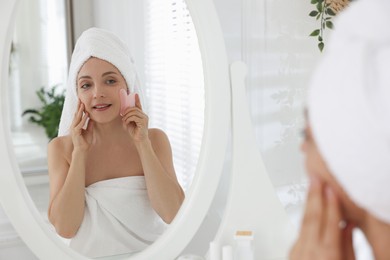 Photo of Smiling woman doing facial self massage with gua sha tool near mirror in bathroom