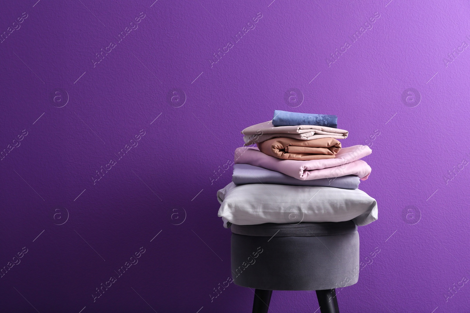 Photo of Stack of clean bed sheets and pillow on stool against purple background. Space for text