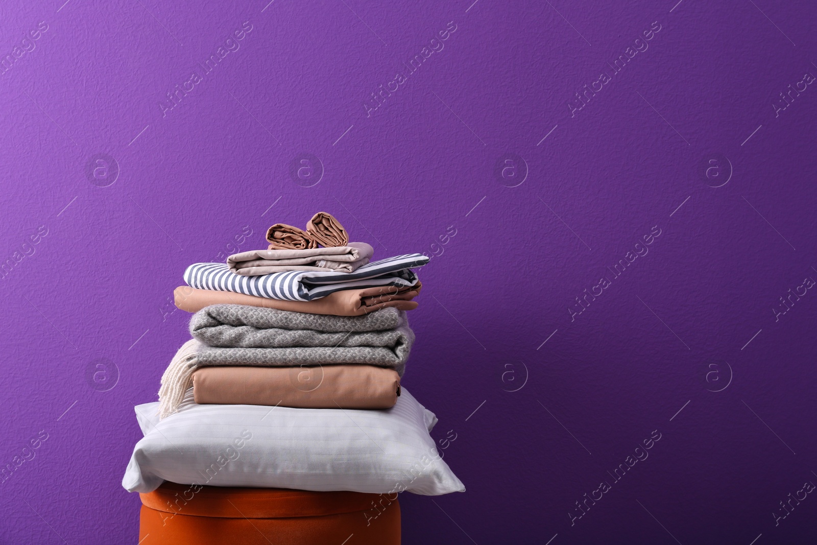 Photo of Stack of clean bed sheets and pillow on stool against purple background. Space for text