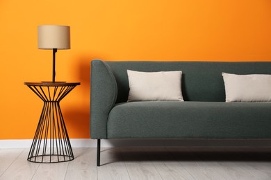 Photo of Stylish sofa with cushions and lamp on side table near orange wall