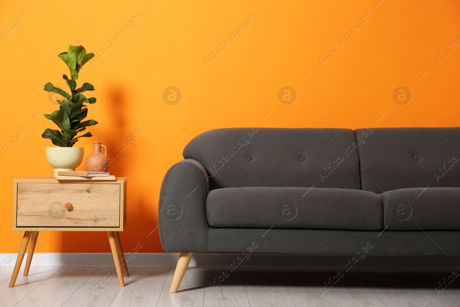 Photo of Stylish sofa with plant and books on side table near orange wall