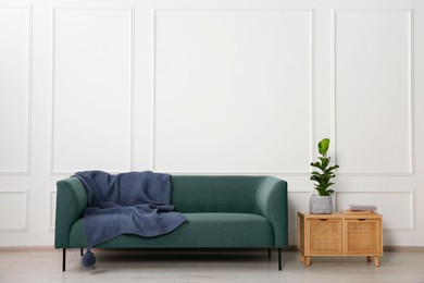 Photo of Stylish sofa with blanket, plant and books on side table near white wall