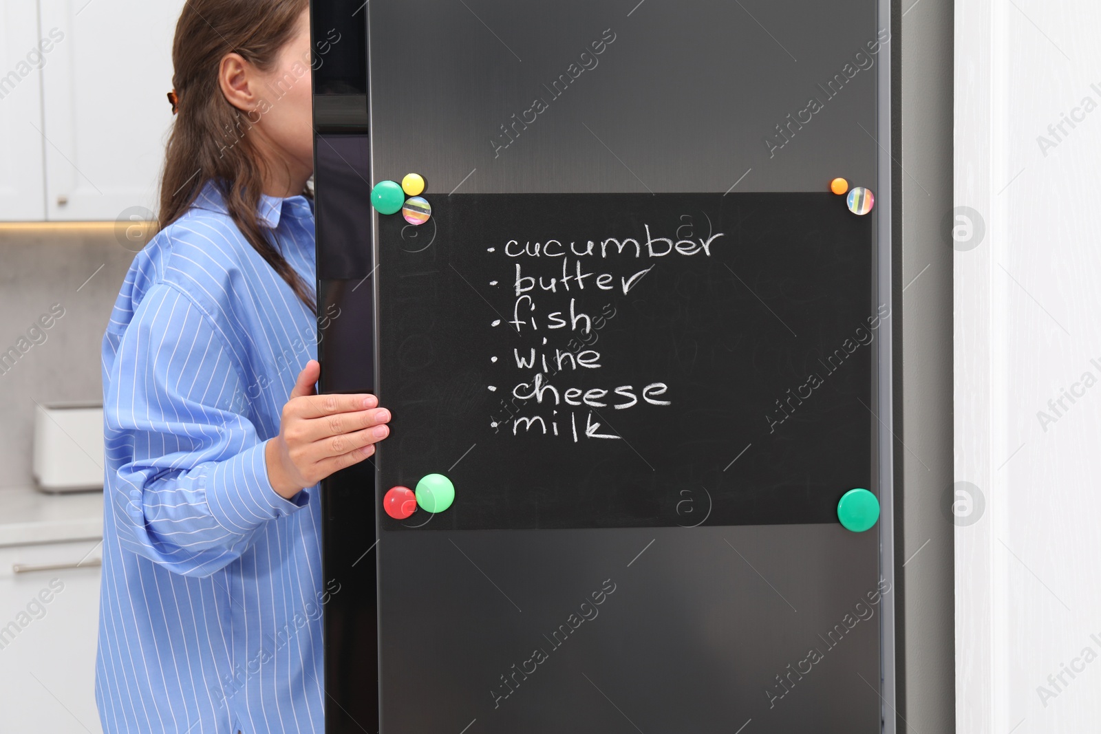 Photo of Woman opening refrigerator with shopping list on magnetic board in kitchen, closeup