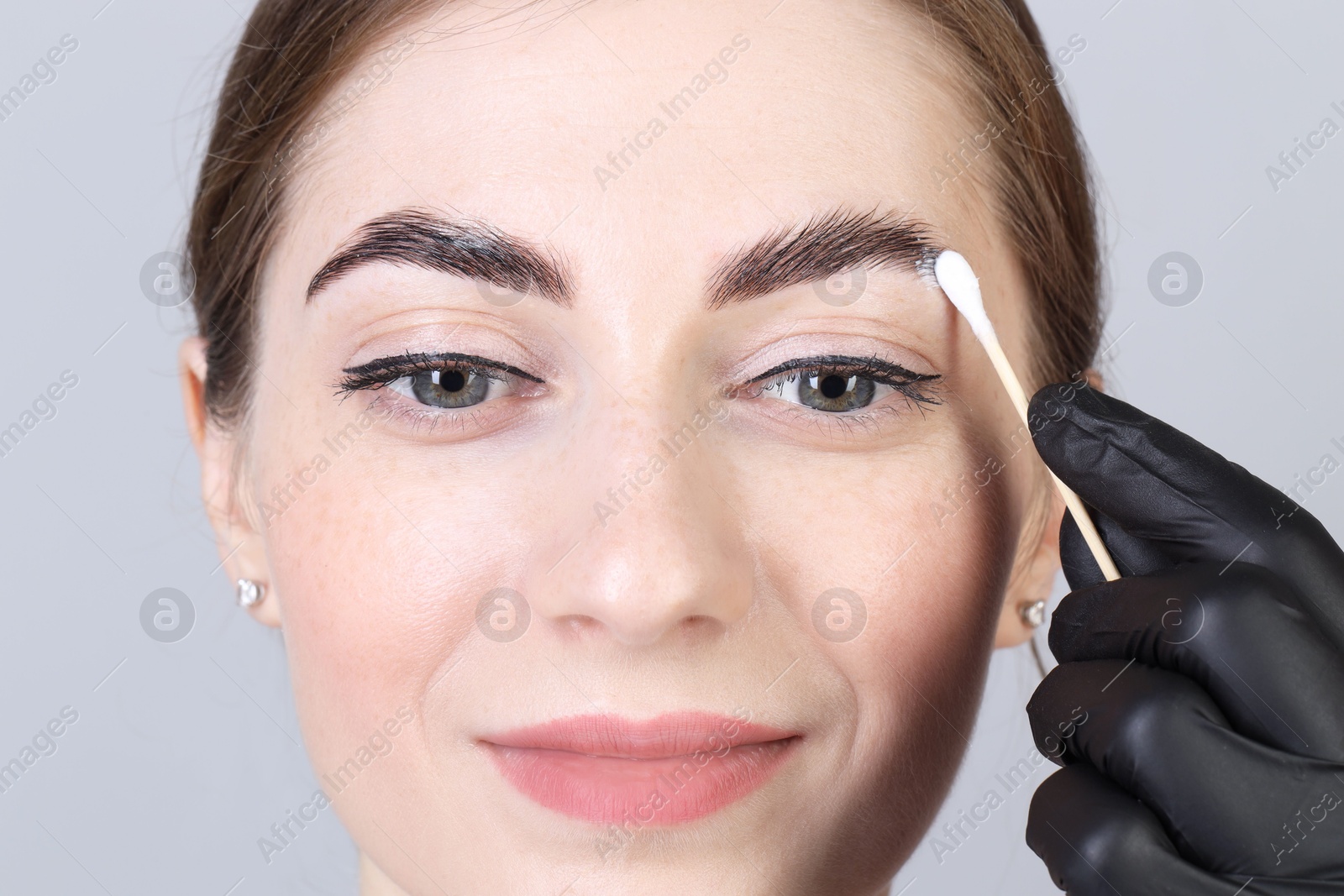 Photo of Brow lamination. Cosmetologist applying cream onto woman's eyebrows against grey background, closeup