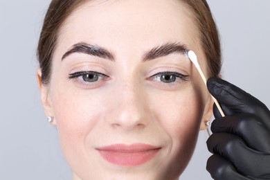 Photo of Brow lamination. Cosmetologist applying cream onto woman's eyebrows against grey background, closeup