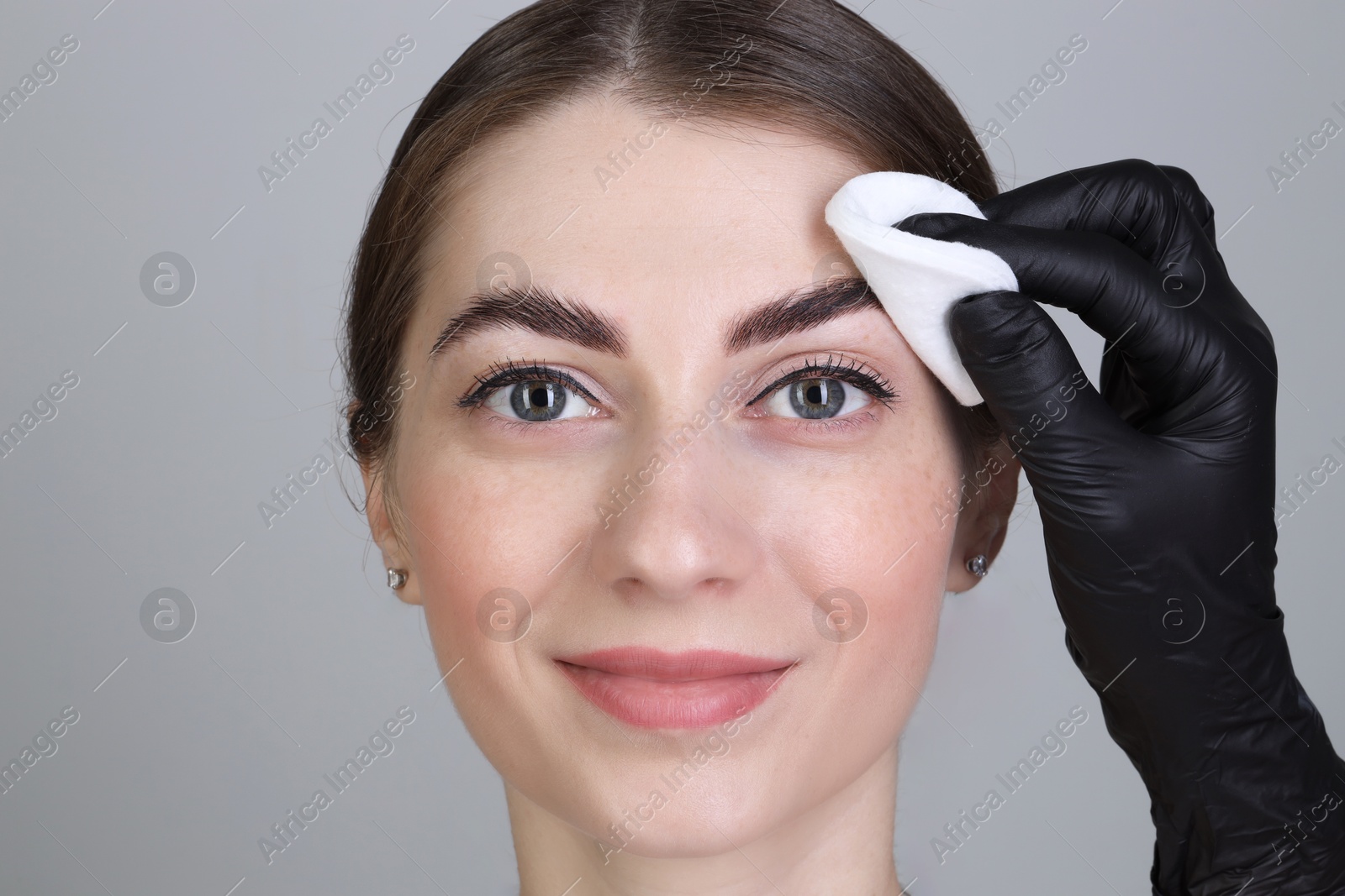 Photo of Brow lamination. Cosmetologist wiping woman's eyebrows with cotton pad against grey background, closeup