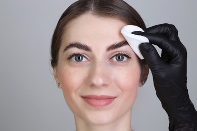 Brow lamination. Cosmetologist wiping woman's eyebrows with cotton pad against grey background, closeup