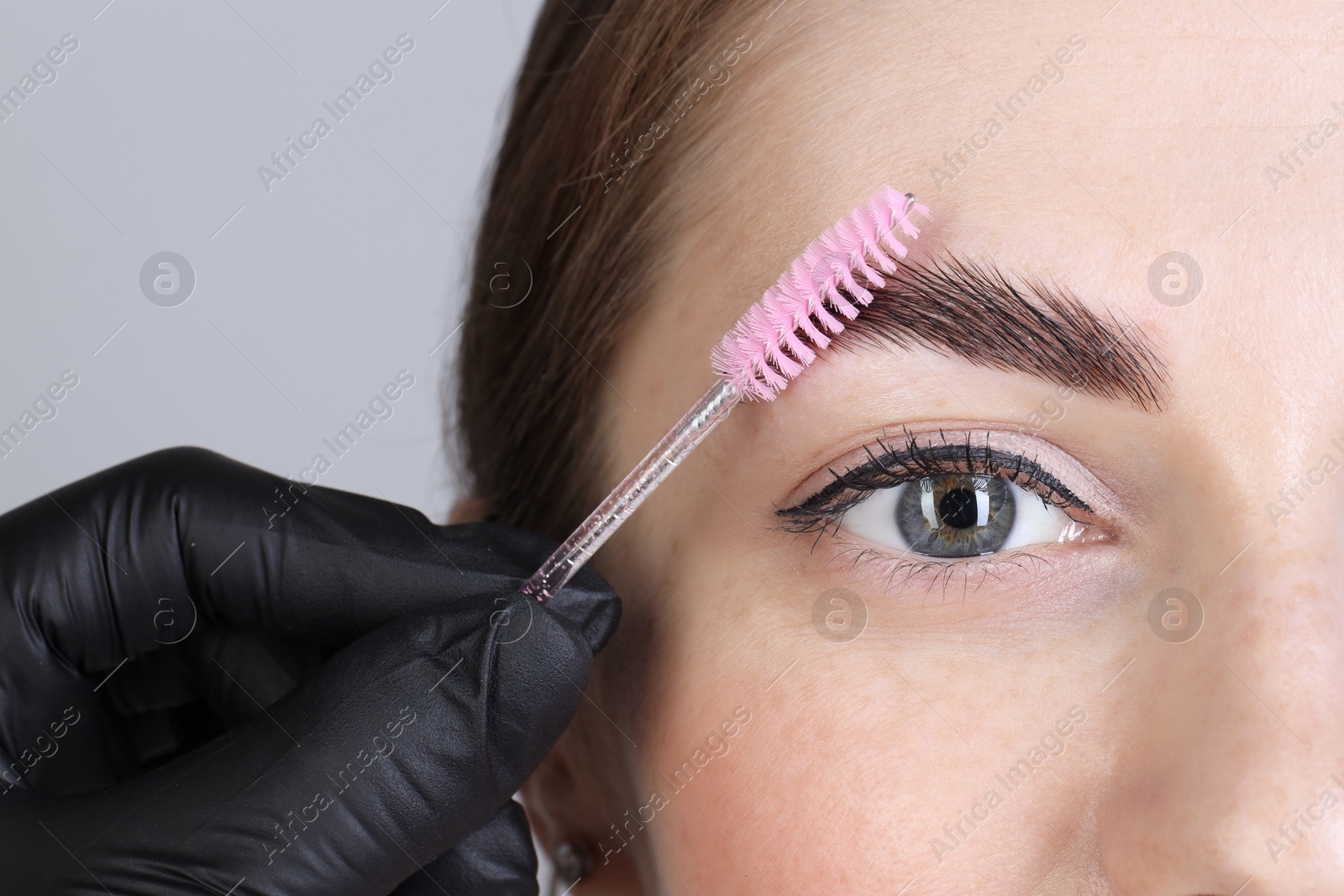 Photo of Brow lamination. Cosmetologist combing woman's eyebrows with brush against grey background, closeup