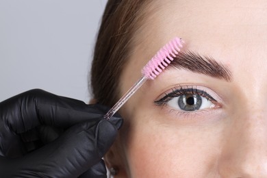 Brow lamination. Cosmetologist combing woman's eyebrows with brush against grey background, closeup