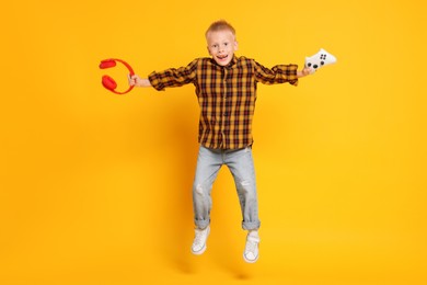 Happy little boy with controller and headphones jumping on orange background