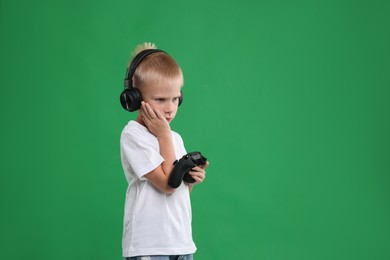 Photo of Upset little boy in headphones with controller on green background. Space for text