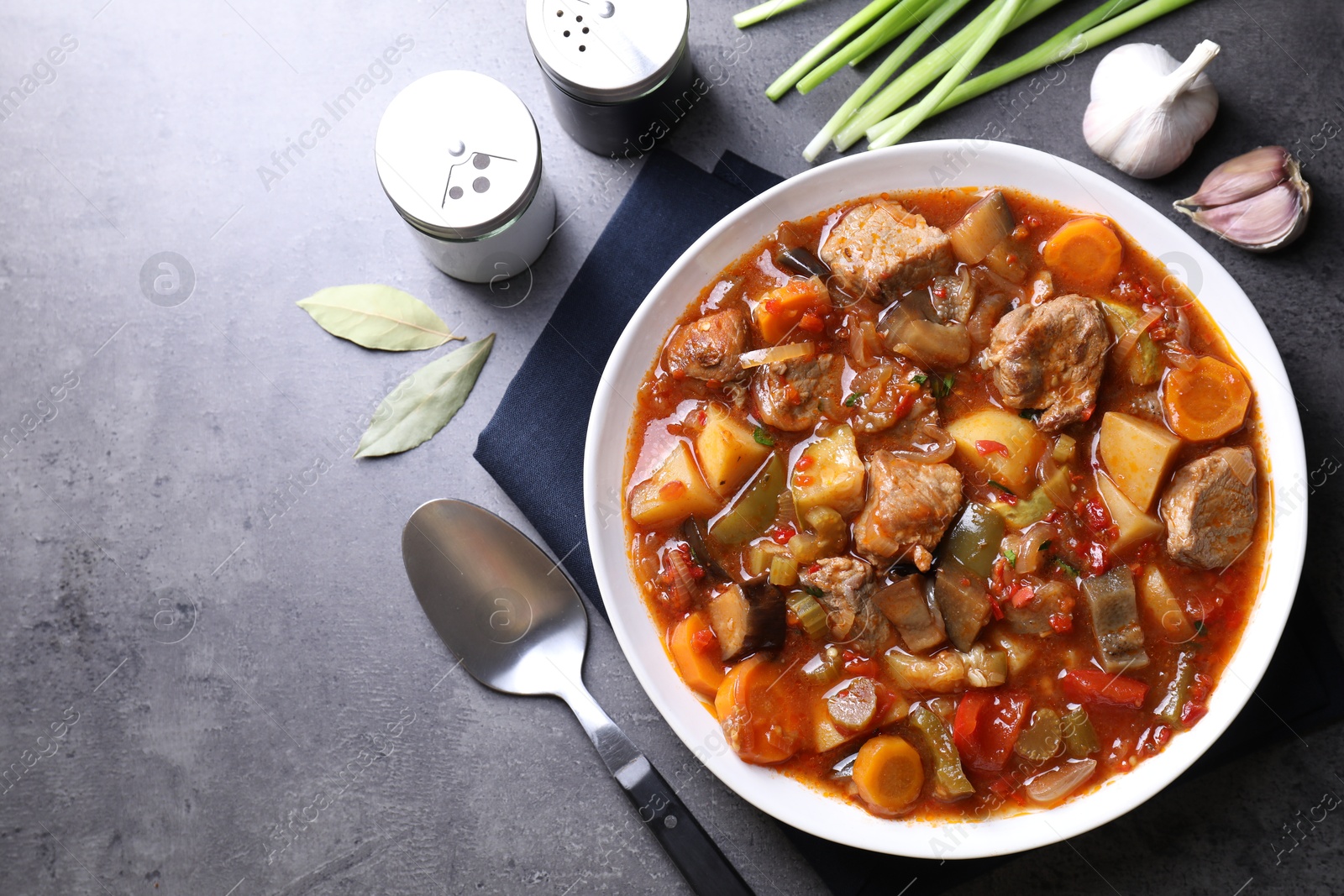 Photo of Delicious stew with vegetables in bowl, spoon and spices on grey table, flat lay