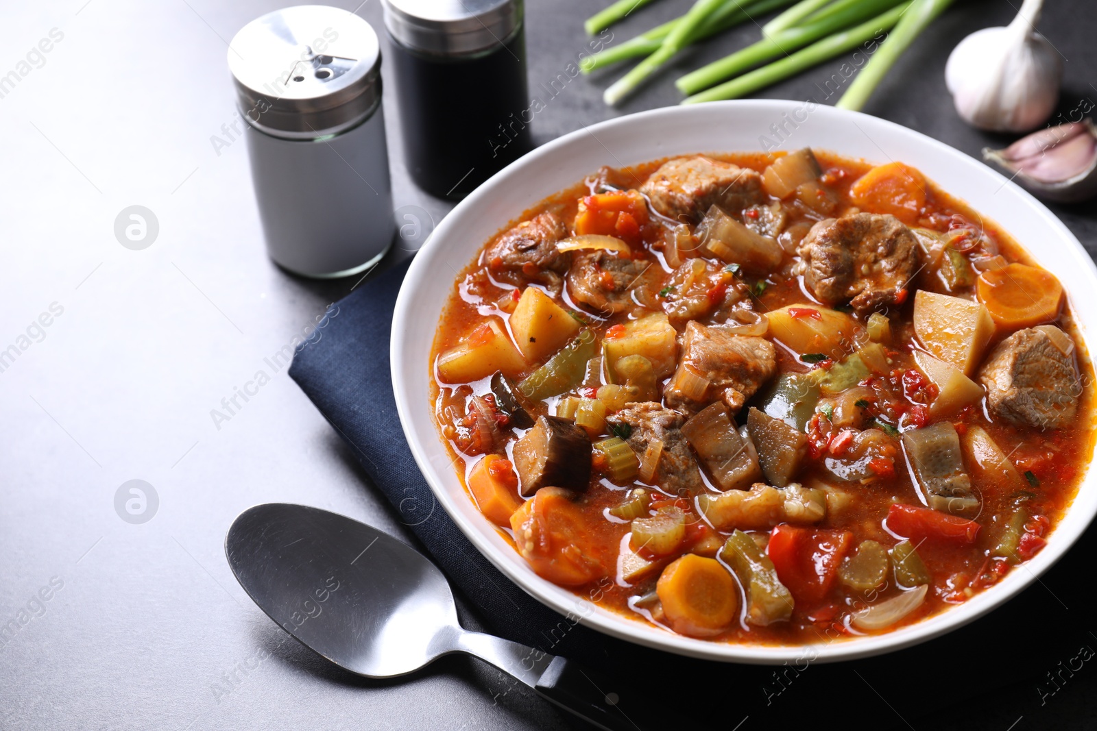 Photo of Delicious stew with vegetables in bowl, spoon and spices on grey table, closeup