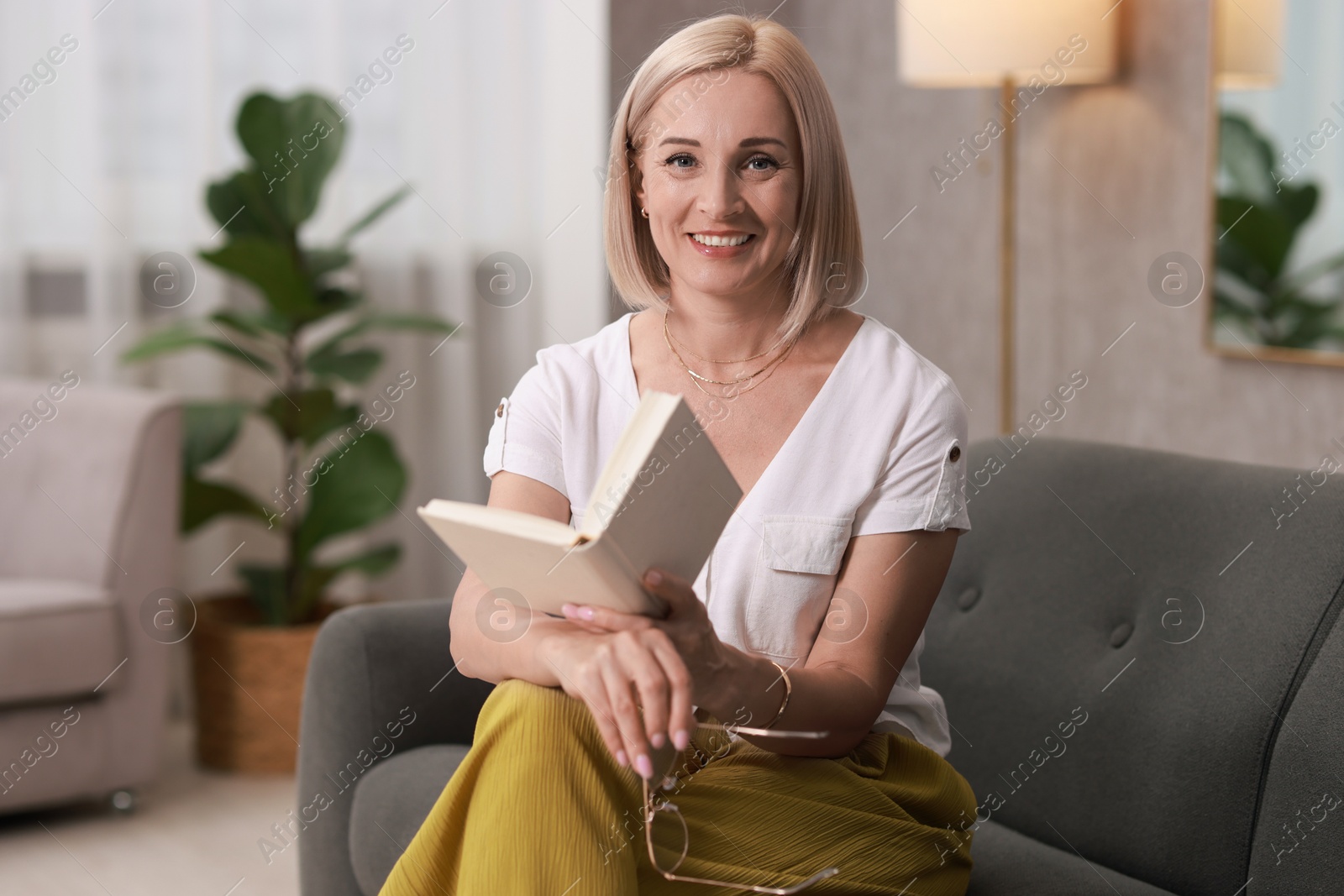 Photo of Portrait of smiling middle aged woman with glasses reading book on sofa at home